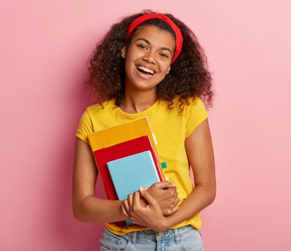Curly lovely college student has positive expression, prepares for exam and reads literature, wears yellow t shirt, poses over pink background. Good emotions, ethnicity and studying concept.