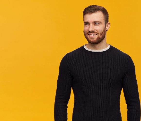 Smiling handsome young man with beard in black longsleeve standing and looking away to the side over yellow background Smiling handsome young man with beard in black longsleeve standing and looking away to the side over yellow background